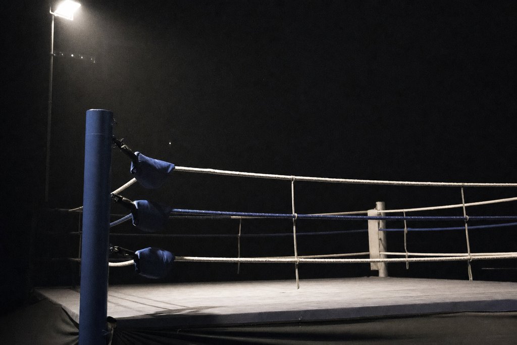 Leerer, ausgeleuchteter Boxring in einer dunklen Halle, im Vordergrund der blaue Eckpfosten